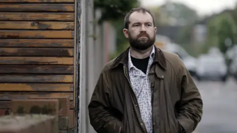 The BBC's undercover reporter, Max, a man with brown hair and a beard, walking down the street with his hands in the pockets of his jacket