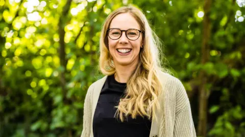 Cassandra Lane A blonde woman with long hair over her shoulder, smiles at the camera wearing round glasses in front of some trees