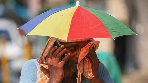 Getty Images A vendor with an umbrella over his head talks on mobile phone as he wipes his face with a cloth on a hot summer day in Varanasi on May 27, 2024