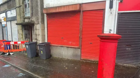 The former Belville Street Post Office  - a small shop with red shutters pulled down