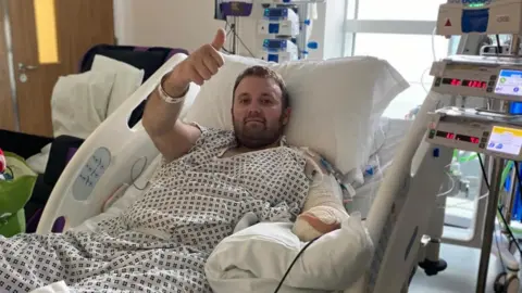 John Knight A picture of Jason taking in hospital. He is wearing a hospital gown and is smiling and giving a thumbs-up to the camera. His is attached to several different machines and his amputated left arm is shown heavily bandaged. 