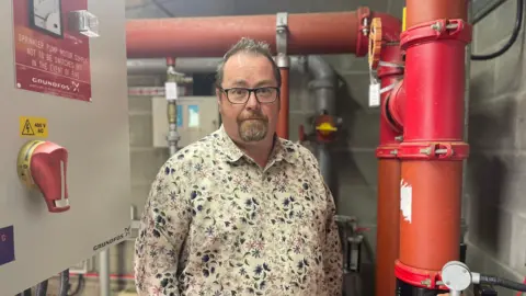 BBC Head teacher Gareth Lewis standing in front of the school sprinkler system