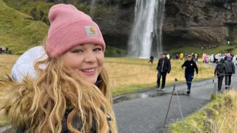 Marcus Handstock A young woman wearing a pink woollen hat, looks over her shoulder and smiles into camera in front of a waterfall