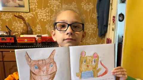 Clarke/BBC News A white girl with black-framed glasses holds up a workbook. She has drawn a brown cat on the left page and a yellow mouse with a red tail on the right page. In the background is a red model train set and there are yellow walls.