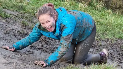 Kim/BBC A white girl in a blue waterproof jacket kneels in the mud, smiling, mud on her face and in her hair 